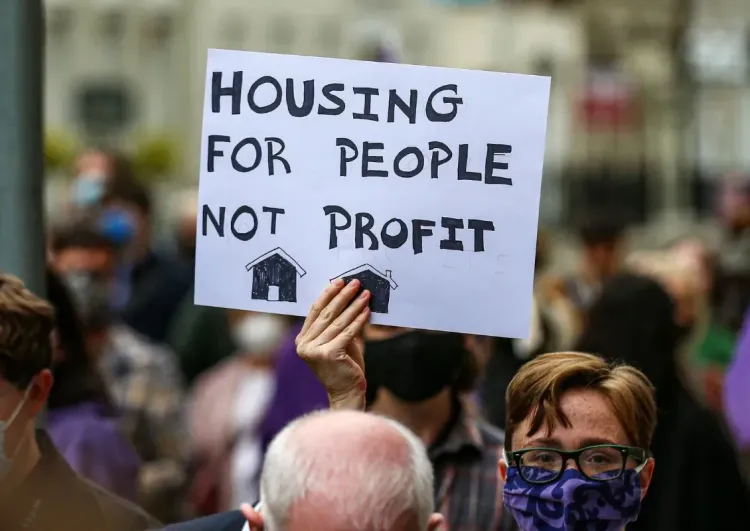 Photo of a young person in a crowd holding up a placard of black text over a white background reading: "Housing for People; Not Profit".